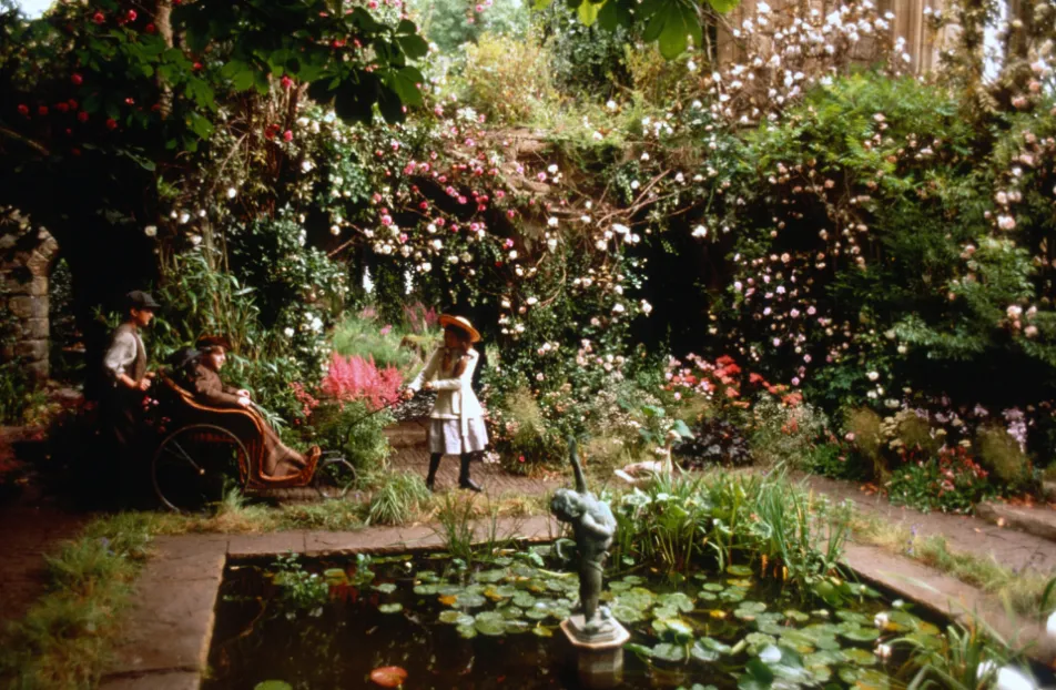 Still from The Secret Garden (1993): Three children- Mary, Colin, and Dickon- entering an area in the titular secret garden. Dickon is pulling Colin's wicker wheelchair and Mary is energetically pulling it. The walled outdoor room is lush and overgrown with vines and flowers. The floor is cobblestone and contains a rectangular lily pond.